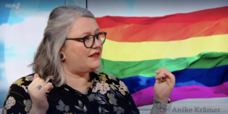 Anike Krämer schaut zur Seite und gestikuliert. Im Hintergrund ist eine Regenbogenflagge zu sehen