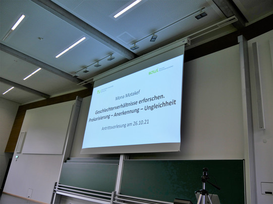 Das Bild zeigt das Titelbild der Präsentation auf der Leinwand bei der Antrittsvorlesung von Prof. Dr. Mona Motakef im Hörsaal.